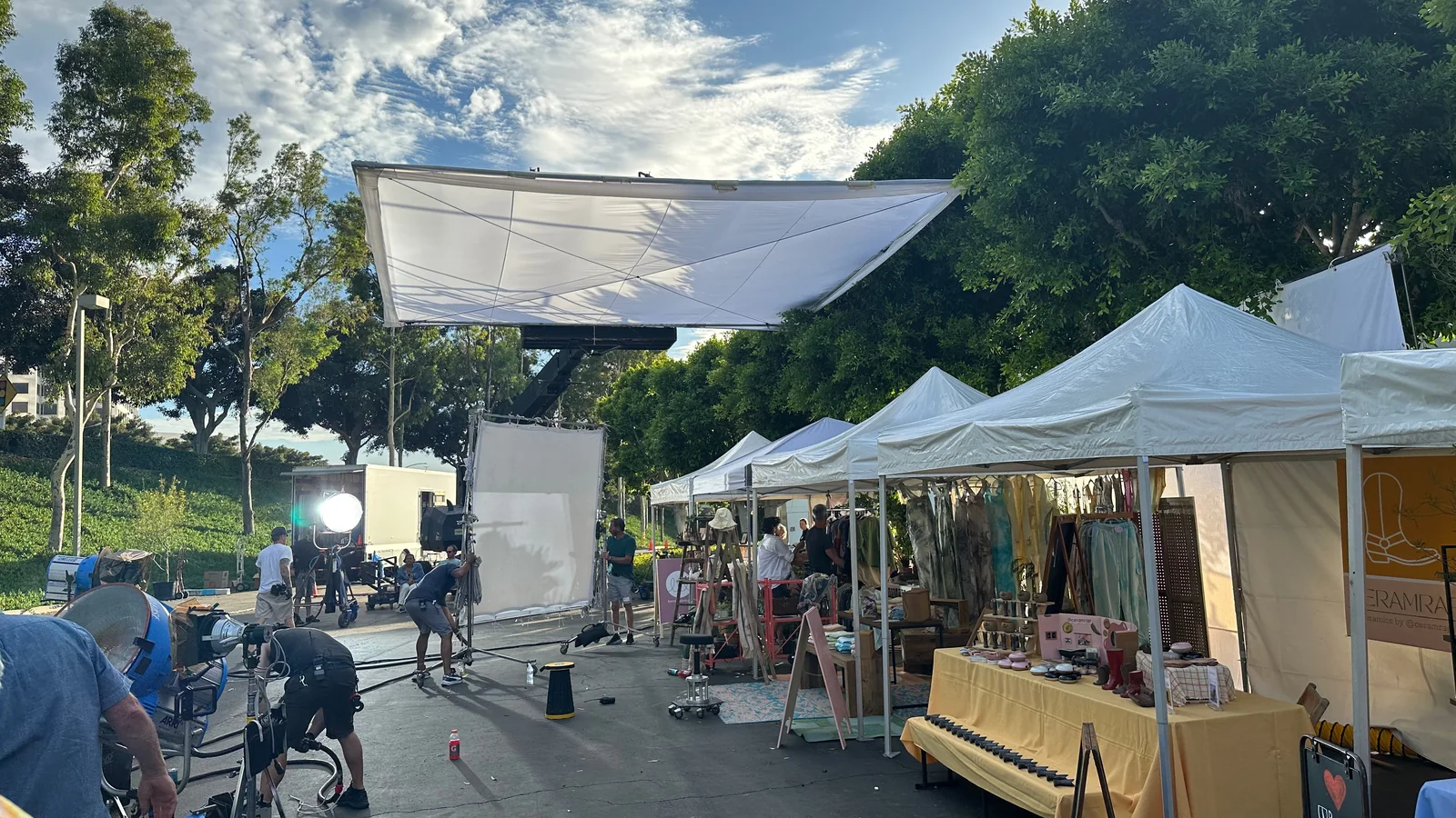A film crew working on an outdoor market set beneath large diffusion, surrounded by tents and lighting.