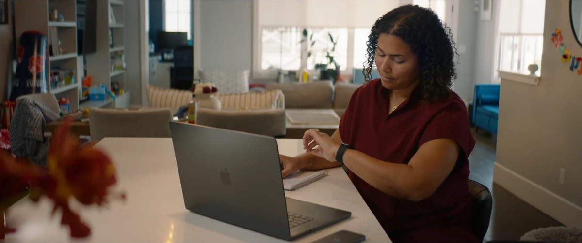 A woman checking her Apple Watch beside a MacBook in the kitchen.