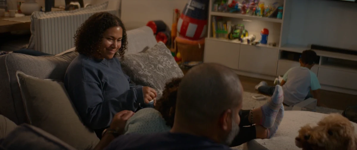 A woman sits with her family in a living room, smiling toward the person beside her.