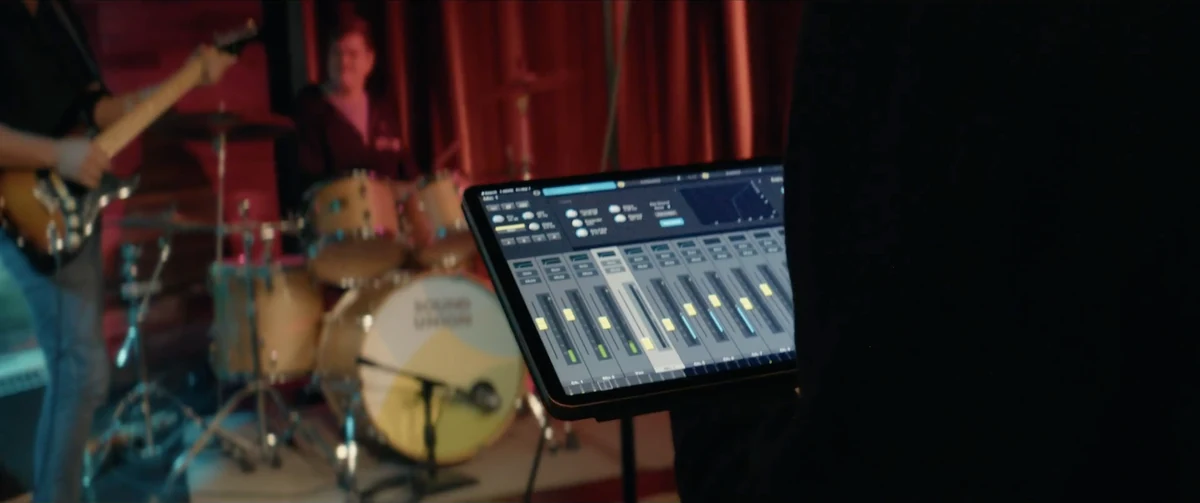 An iPad mixing console sits in front of a drummer and a red stage curtain.