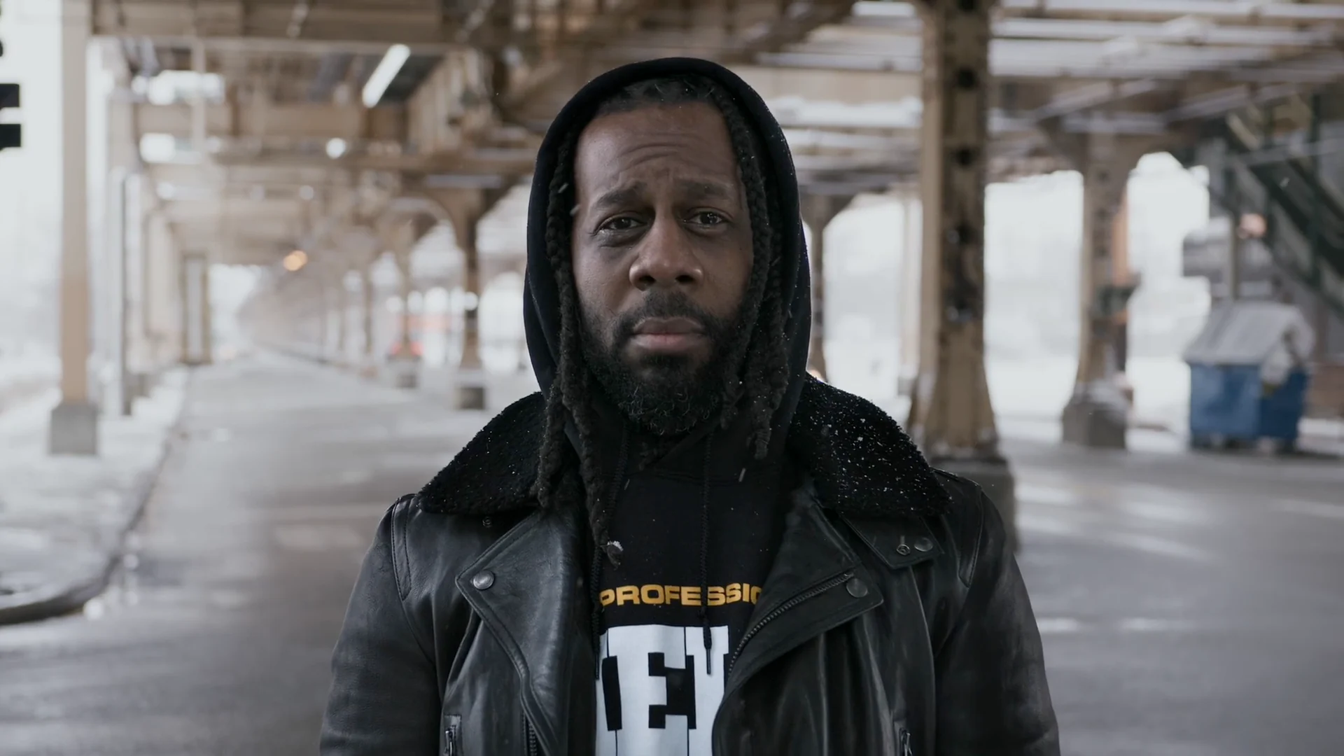 Barber Nas standing beneath elevated train tracks in a frame from Booksy’s Self Made series.
